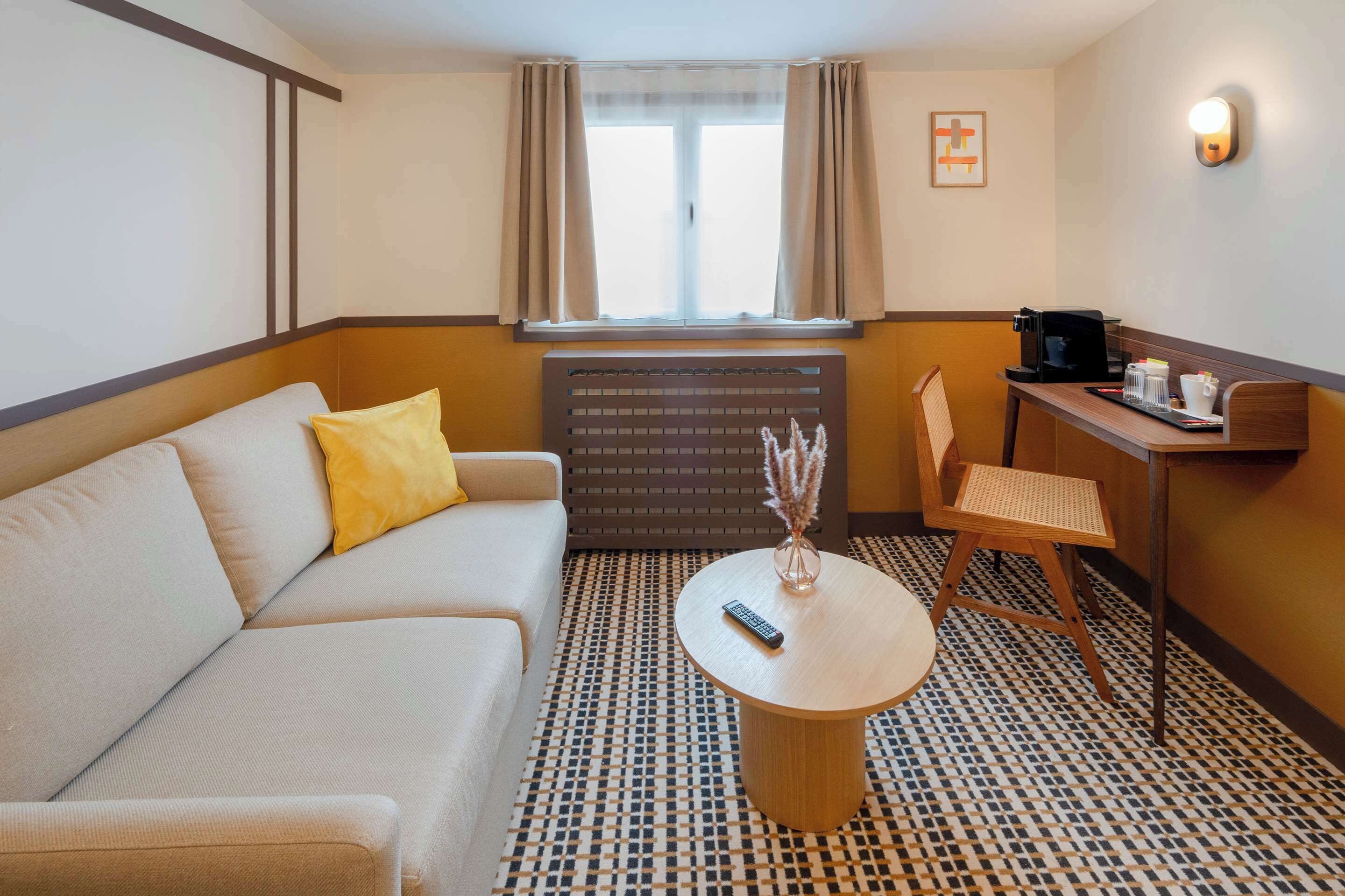 Une chambre d’hôtel avec un canapé, une petite table ronde, une chaise, une machine à café et une fenêtre avec des rideaux.
