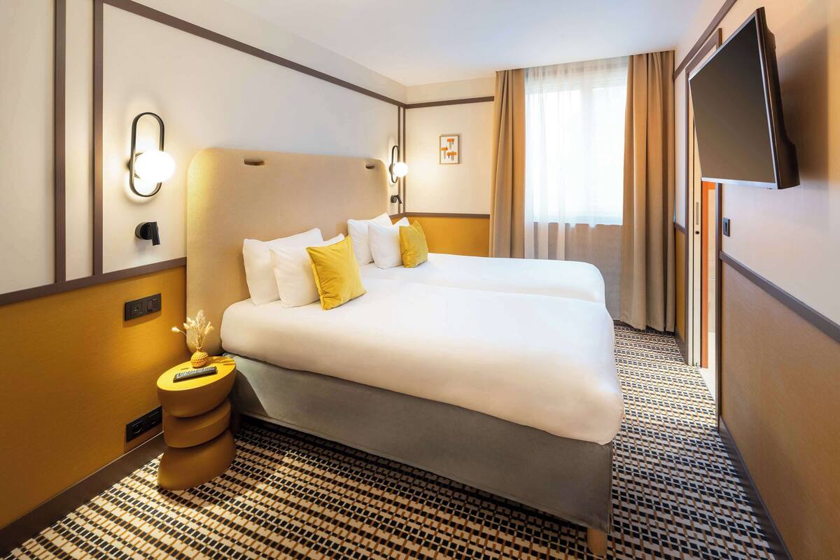 Une chambre d’hôtel avec un grand lit, une table de chevet avec une lampe, un téléviseur à écran plat et une moquette à motifs.