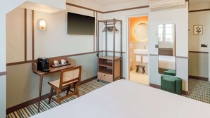 A hotel room with a bed, a desk with a coffee maker, a chair, a coat rack, a mirror, and a bathroom with a sink and toilet.