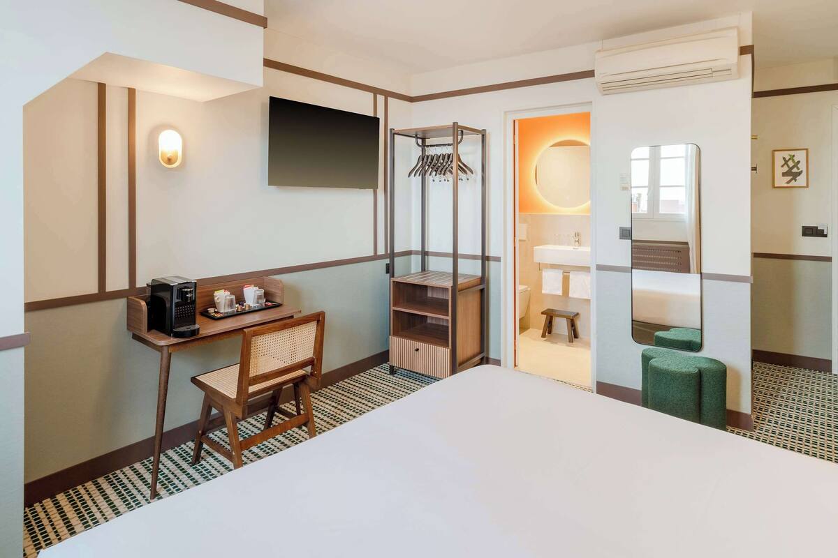 Une chambre d’hôtel comprenant un lit, un bureau avec une machine à café, une chaise, un porte-manteau, un miroir et une salle de bain avec un lavabo et des toilettes.