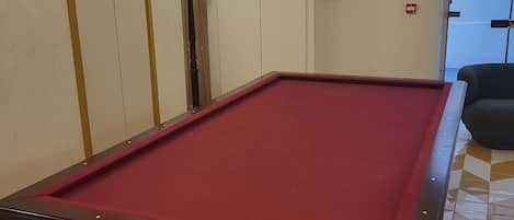 Una mesa de billar con superficie de fieltro rojo, rodeada por un suelo de baldosas estampadas. Hay luces colgantes y una puerta con un cartel en la pared.