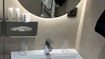 A modern bathroom with a rectangular white sink, two faucets, and a large mirror above it. There are toiletries on a shelf, a towel rack with a towel, and a black bag hanging on the wall.