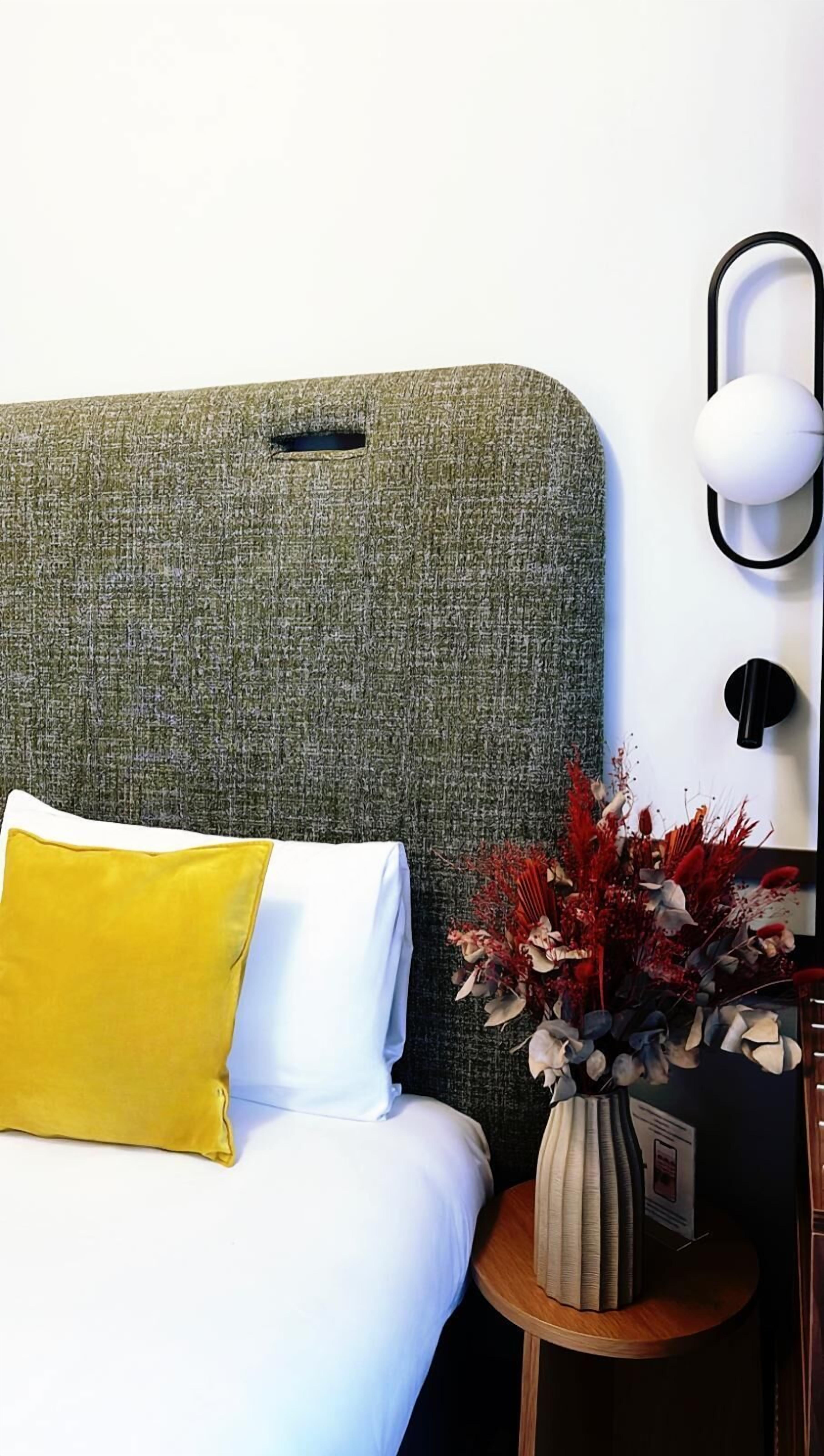 A bed with a green headboard, a white pillow, and a yellow pillow. There is a small wooden nightstand with a vase of red and white flowers.