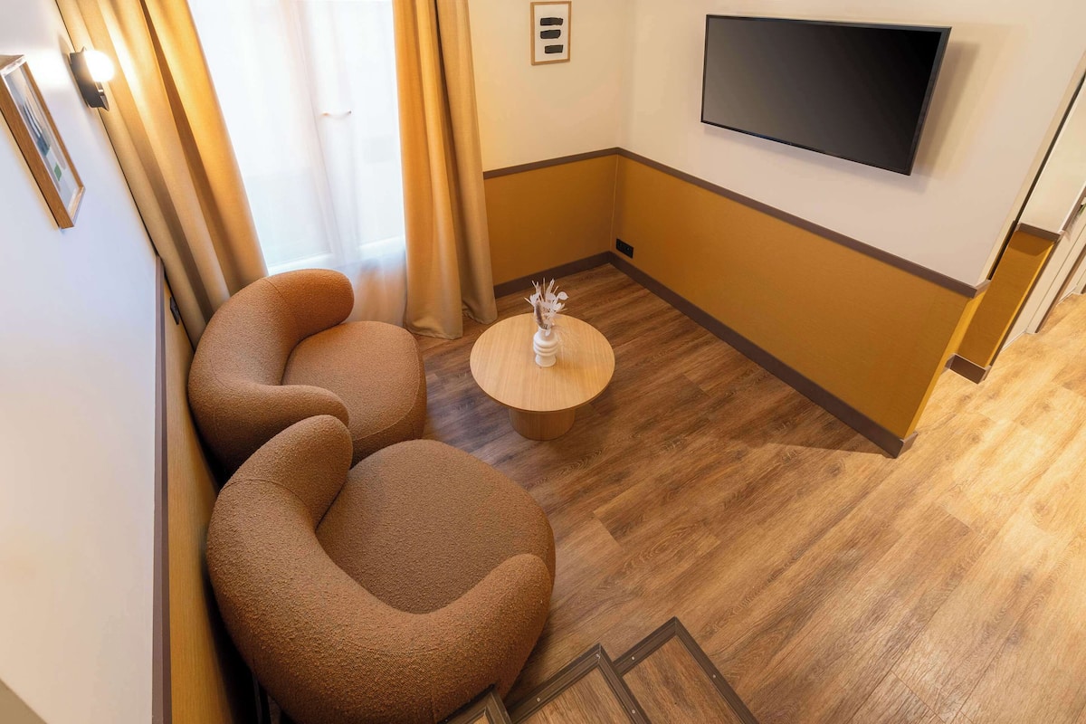 Une chambre d’hôtel dotée d’un téléviseur à écran plat fixé au mur, d’un plancher en bois et de deux fauteuils marron.