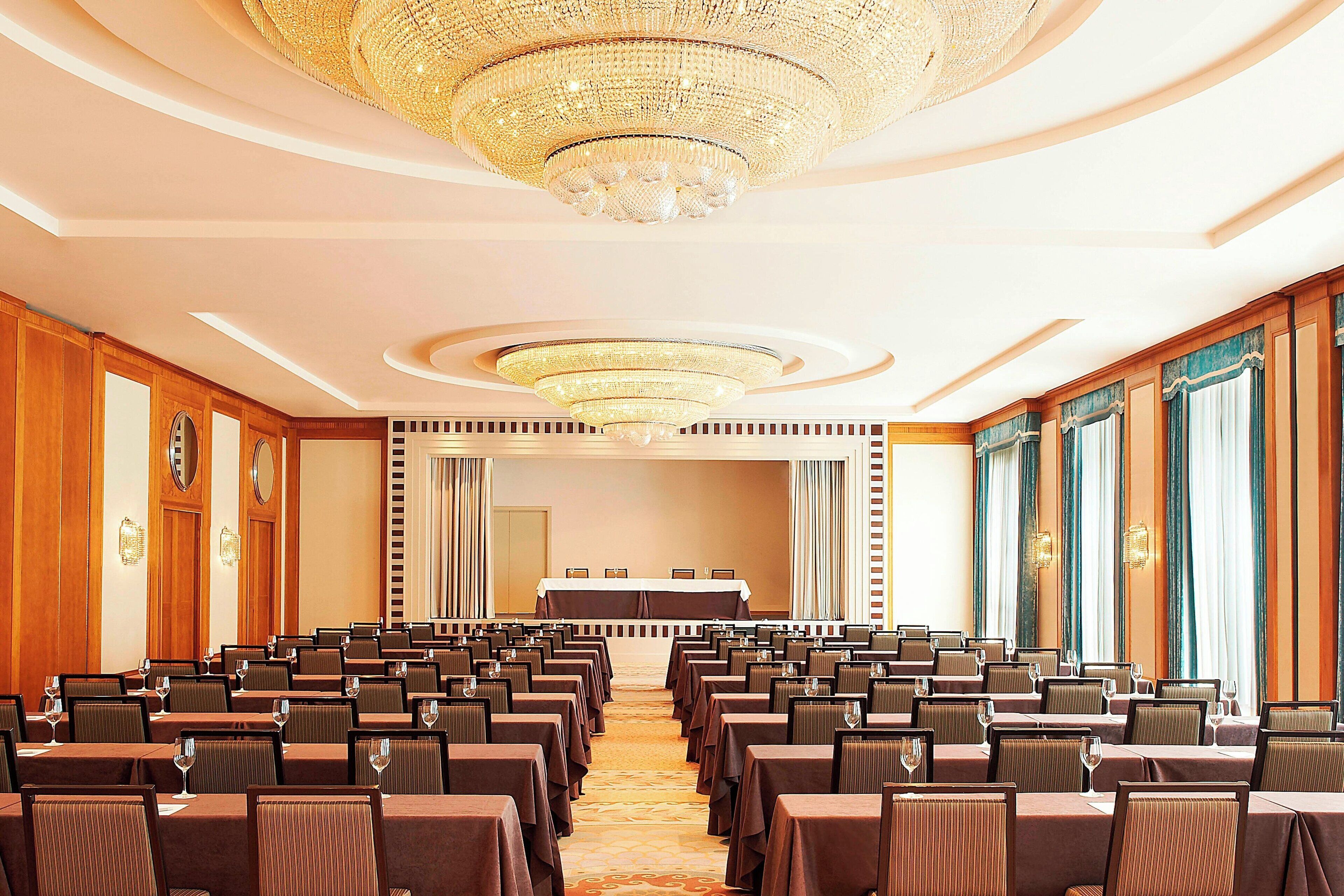 A conference room with rows of tables and chairs, a large chandelier, and a stage with a table and chairs.