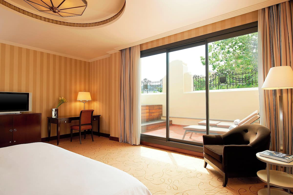 Habitación de hotel con cama, escritorio, silla, televisor, lámpara, mesa y balcón con vista a vegetación.