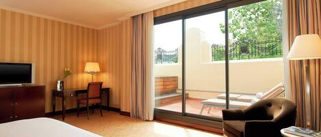 A hotel room with a bed, a desk, a chair, a TV, a lamp, a table, and a balcony with a view of greenery.