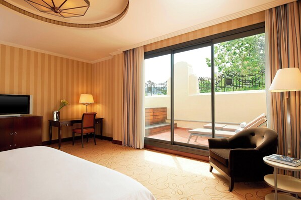 Habitación de hotel con cama, escritorio, silla, televisor, lámpara, mesa y balcón con vista a vegetación.
