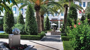 Un patio ajardinado con palmeras, una fuente y un edificio con ventanas grandes al fondo.