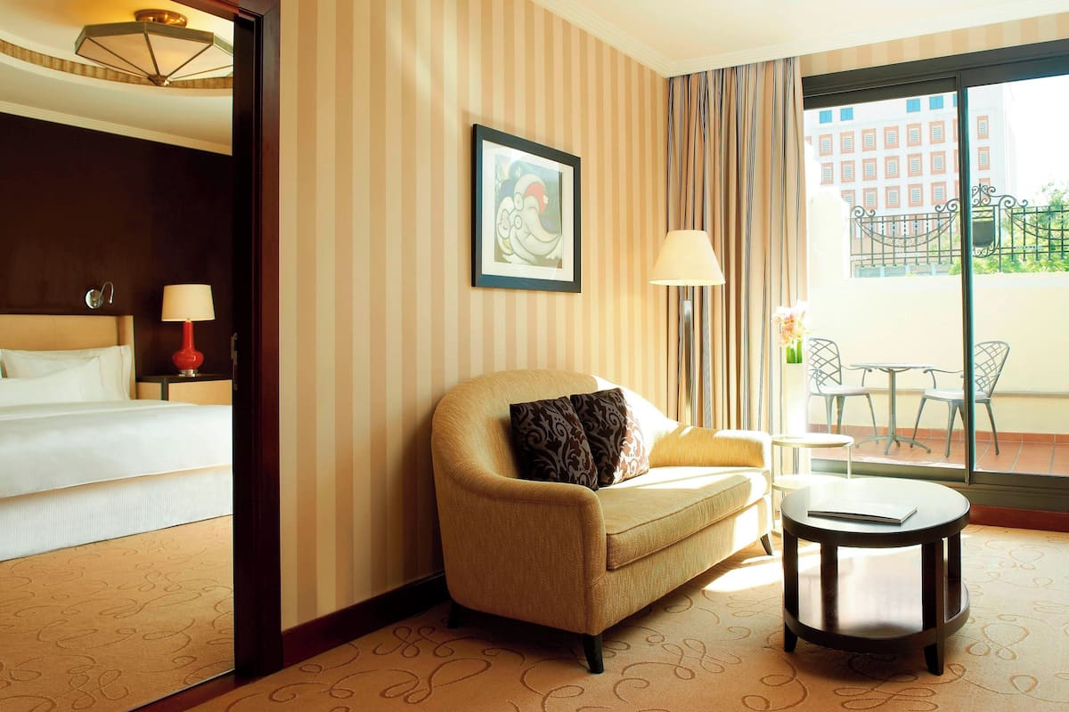 Habitación de hotel con cama, sofá, una mesita y balcón con vistas a edificios.
