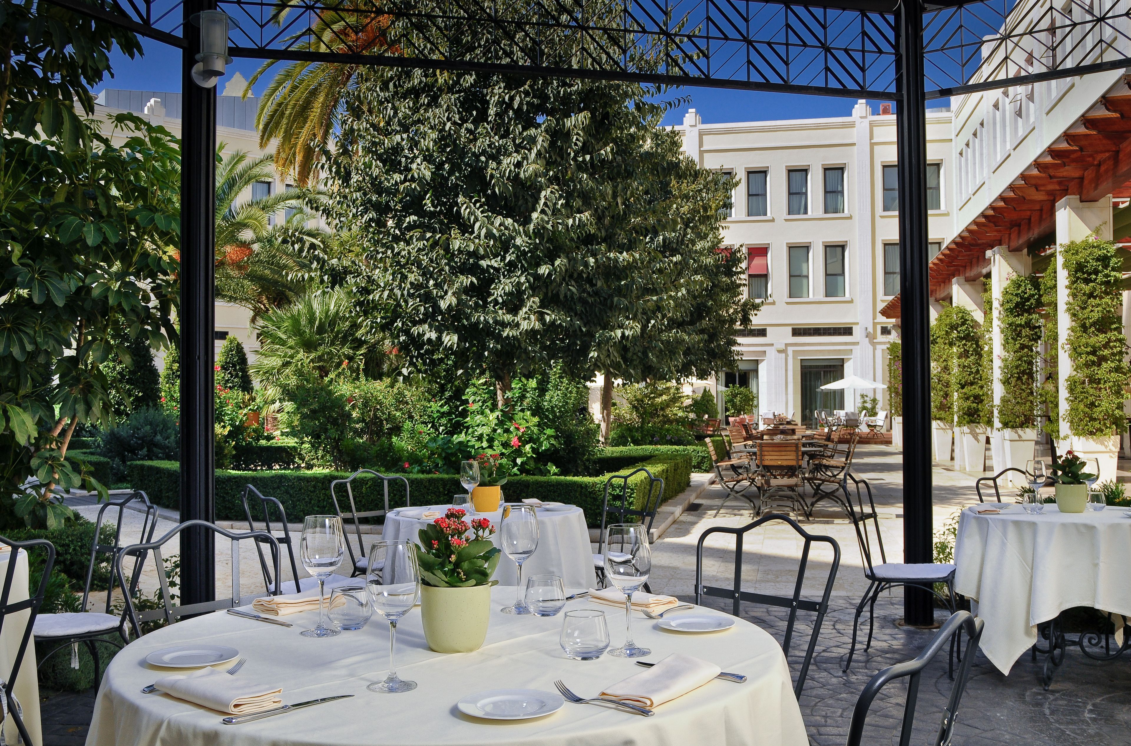 An outdoor dining area with tables set for two, surrounded by greenery and a building in the background.