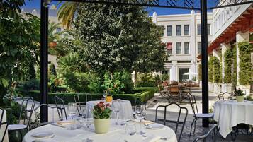 An outdoor dining area with tables set for two, surrounded by greenery and a building in the background.