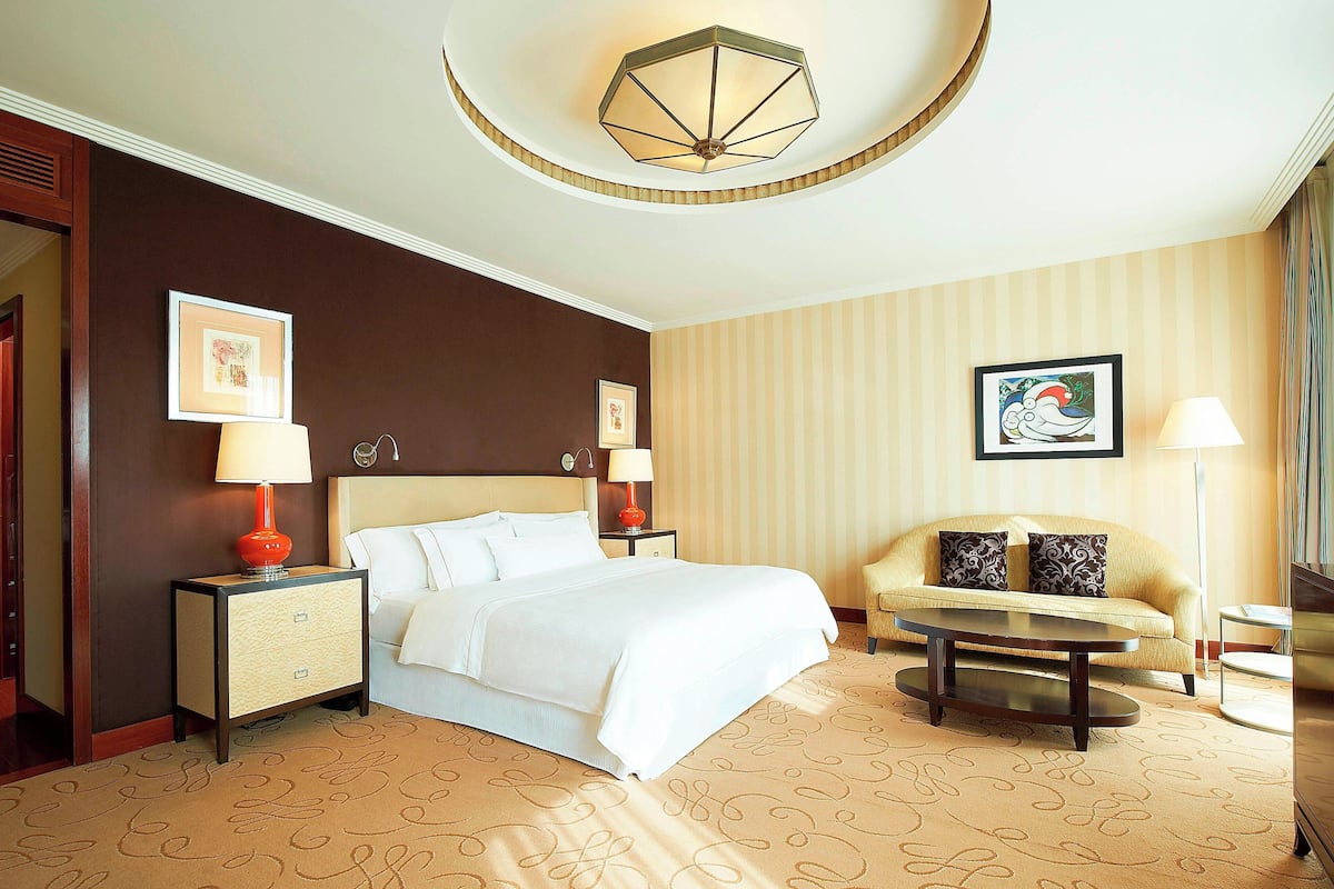 Habitación de hotel con cama, dos mesitas de noche con lámparas, un sofá, una mesa de centro y un candelabro en el techo.