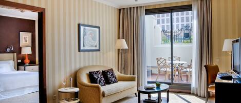 A hotel room with a bed, a sofa, a small table, a TV, and a balcony with a view of buildings.
