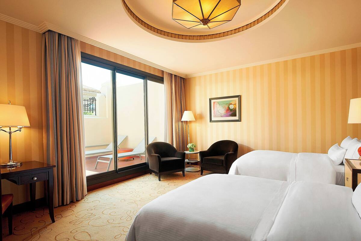 Habitación de hotel con una cama grande, dos sillones, un escritorio, una lámpara, una mesita con una planta y un ventanal con cortinas.