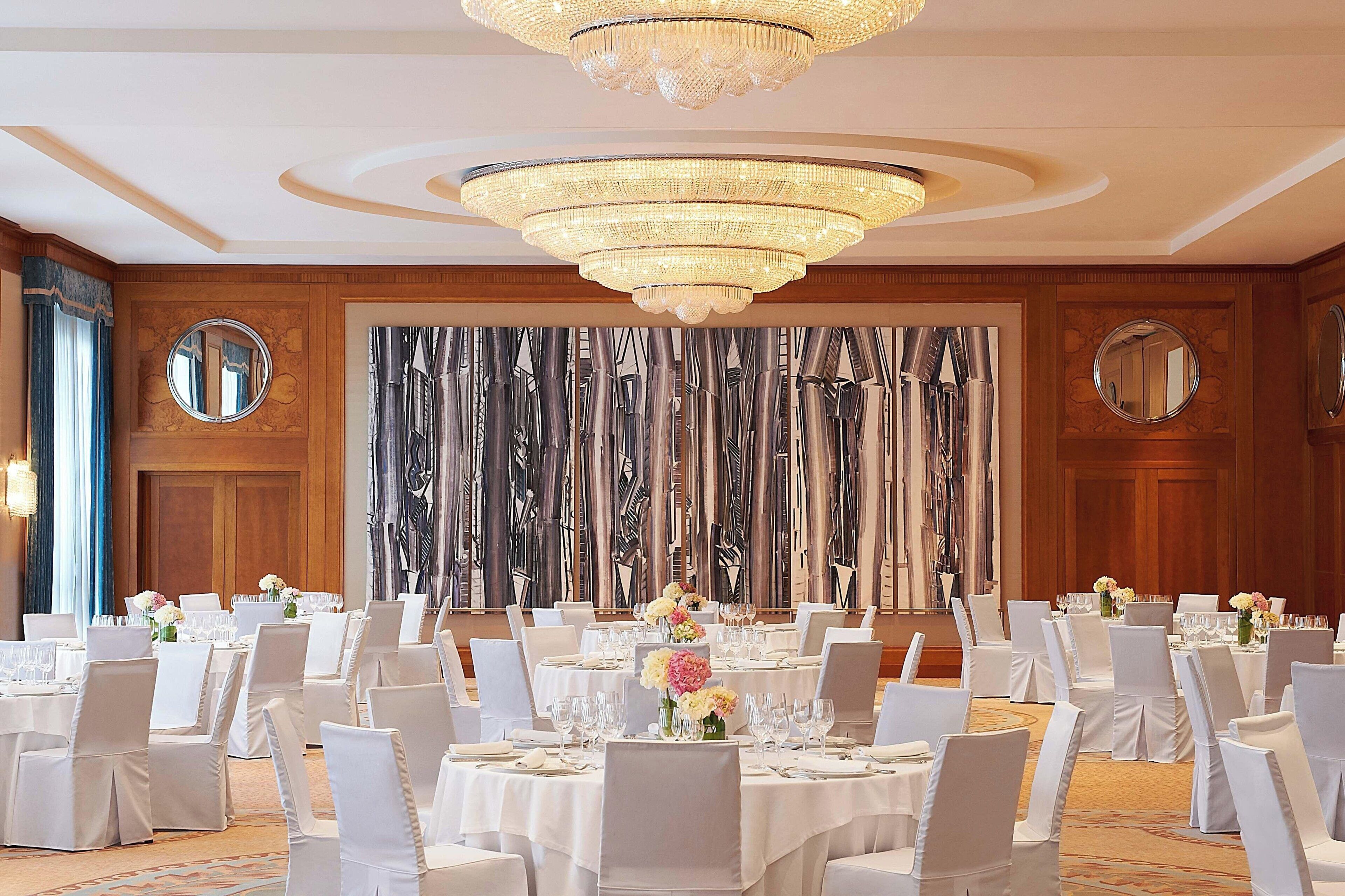 A banquet hall with round tables, white chairs, and chandeliers.