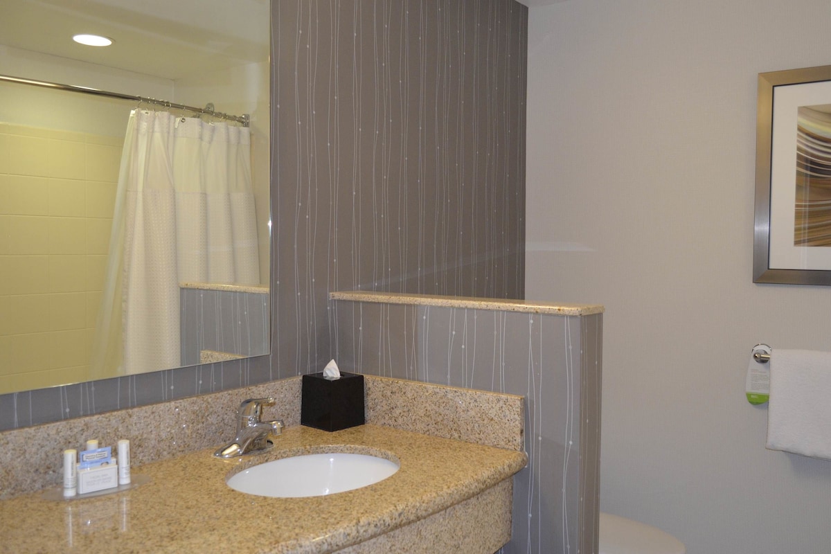 A bathroom with a granite countertop sink, a mirror, a towel dispenser, and a framed picture on the wall.