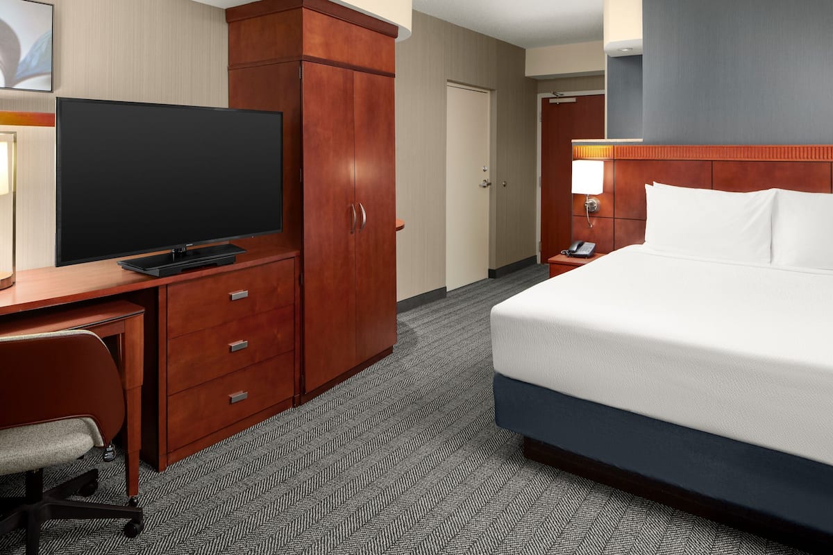 A hotel room with a large flat-screen TV, a wooden wardrobe, a desk with a chair, a bed with white linens, and a nightstand with a lamp.