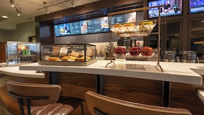A bar area with a display case of baked goods, a fruit basket, and a television screen displaying news. - Courtyard by Marriott Winchester (Winchester)