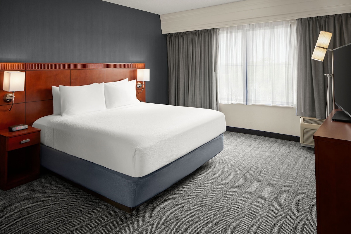 A hotel room with a large bed, wooden headboard, two bedside tables with lamps, a flat-screen TV, and a window with curtains.