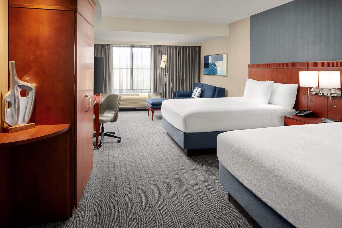A hotel room with two beds, a desk, a chair, a desk lamp, and a large window with curtains.