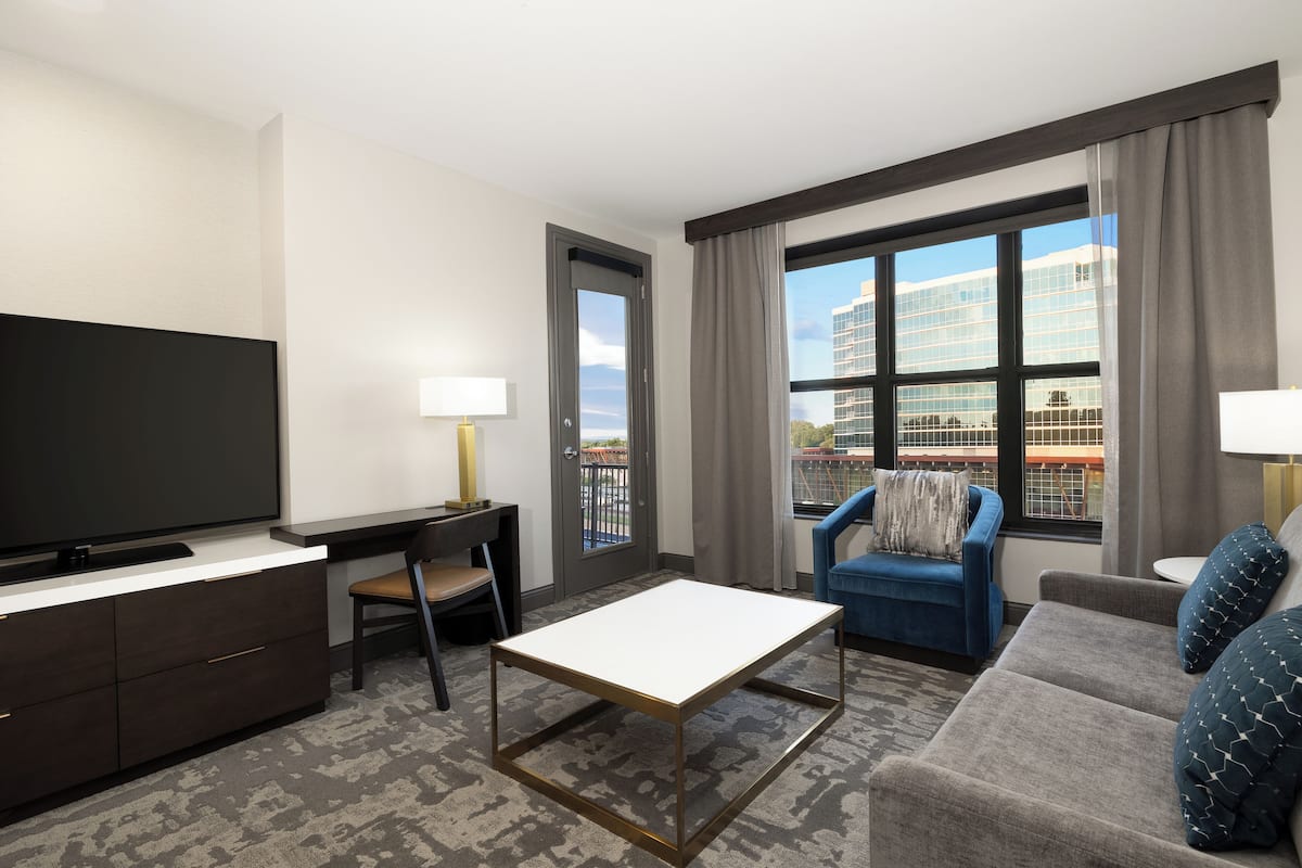 A hotel room with a flat-screen TV, a desk with a lamp, a sofa, a chair, and a large window with a view of buildings.