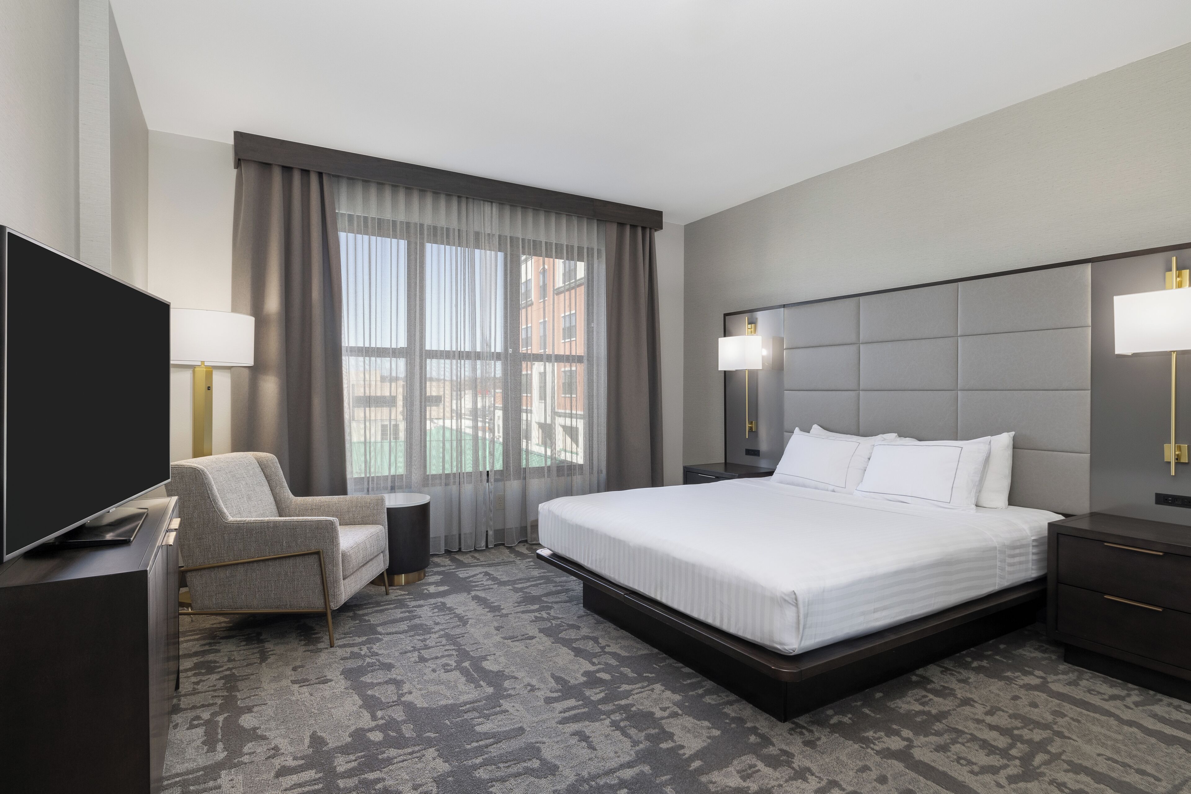 A modern hotel room with a large bed, a flat-screen TV, a comfortable armchair, and a view of the city through large windows.