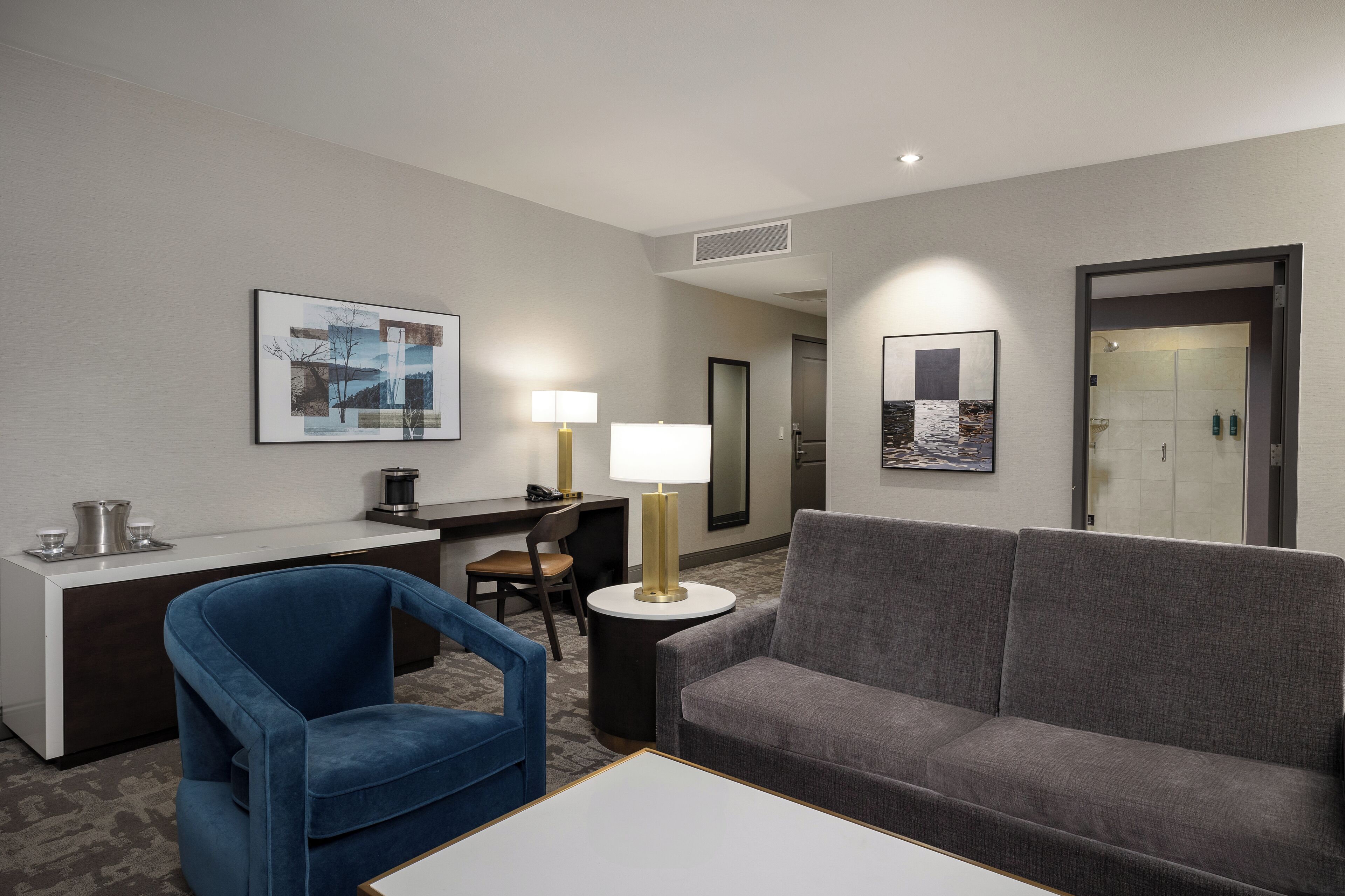 A hotel room with a gray sofa, a blue armchair, a desk with a lamp, and artwork on the walls.