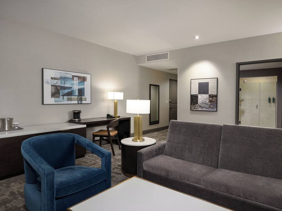 Habitación de hotel con un sofá gris, un sillón azul, un escritorio con lámpara y obras de arte en las paredes.