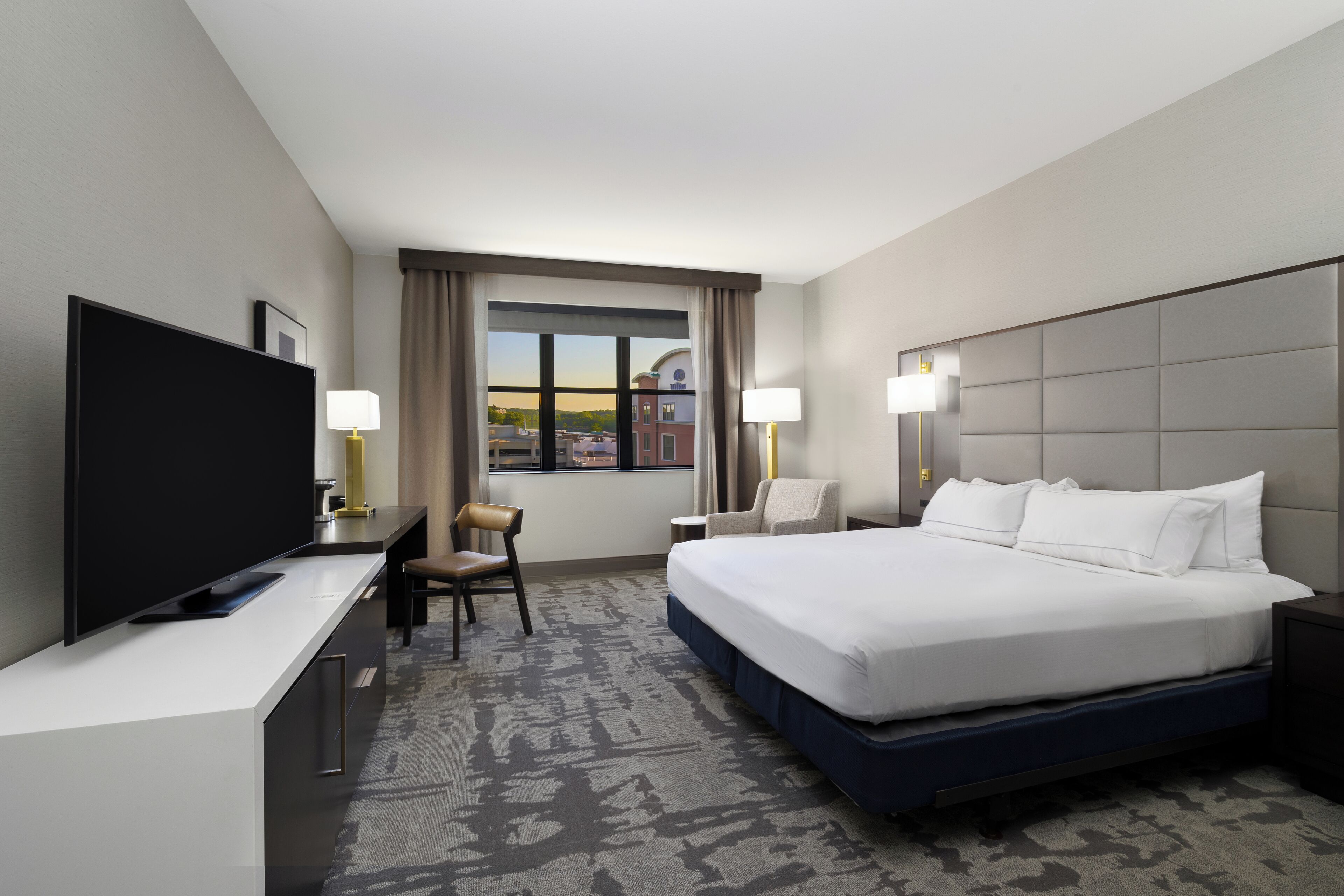 Una habitación de hotel con una cama grande, un escritorio con televisor, dos lámparas, una silla y vista a un paisaje urbano por la ventana.