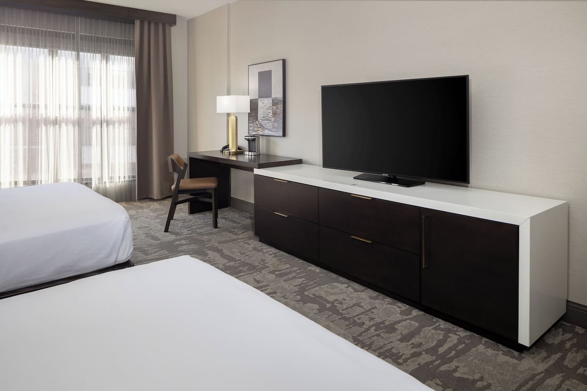 A hotel room with a bed, a desk with a chair, a television, and a large window with curtains.