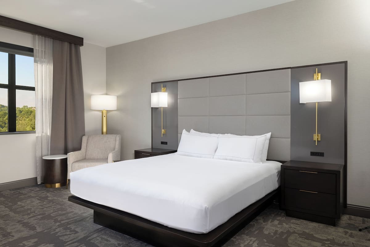 A hotel room with a large bed, two bedside tables with lamps, a chair, and a view of greenery through the window.