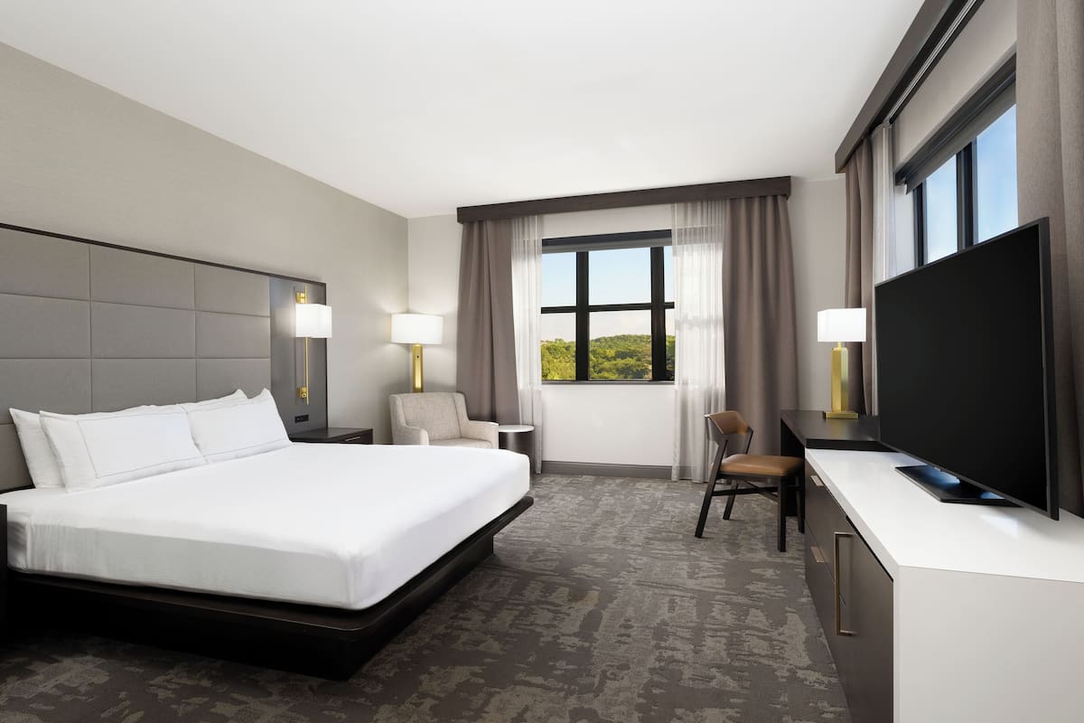 A modern hotel room with a large bed, a desk with a flat-screen TV, a chair, and a window with curtains.
