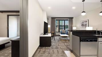 A modern hotel room with a kitchenette, a flat-screen TV, a sofa, and a dining area.