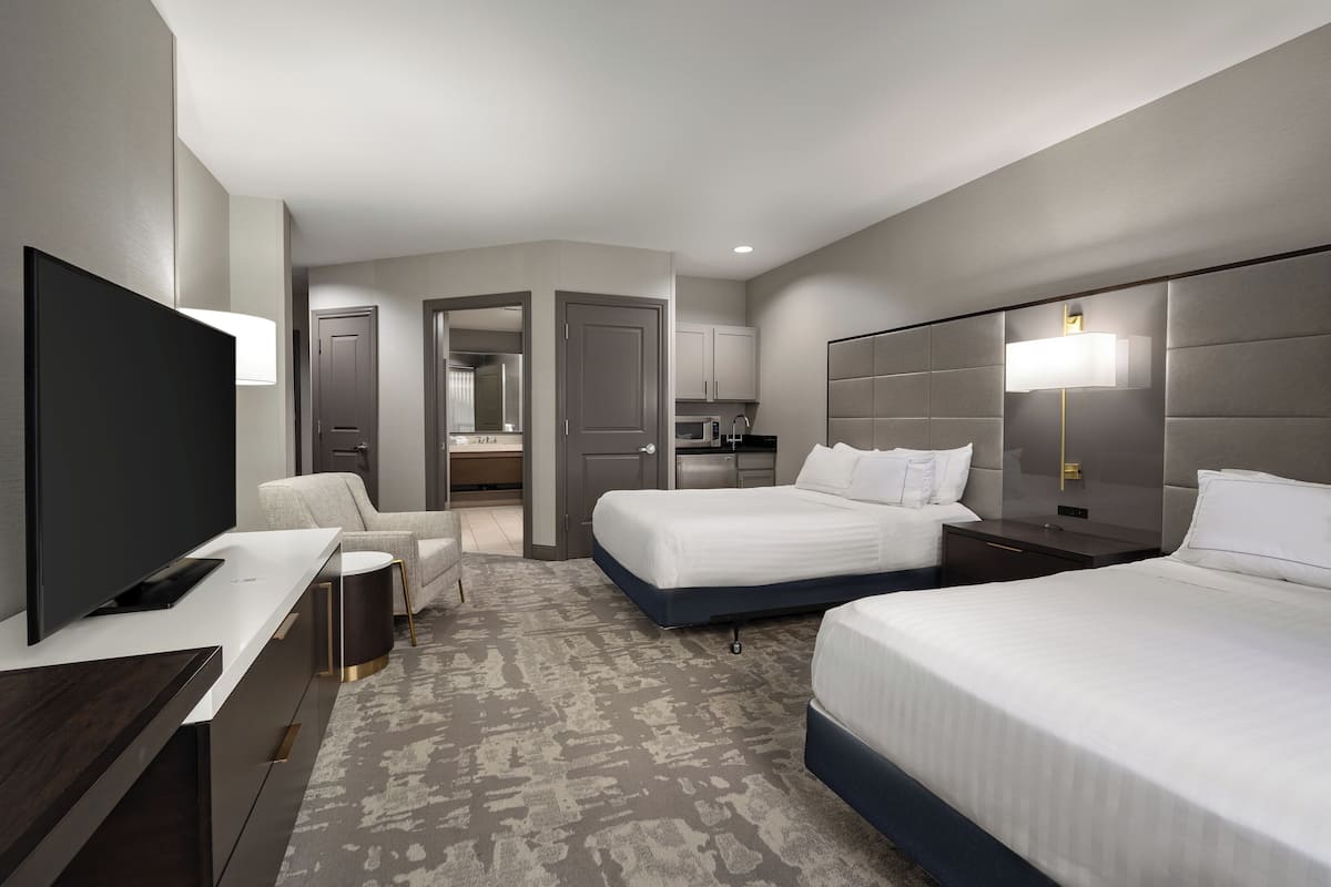 A modern hotel room with a flat-screen TV, two beds, a desk, a chair, a small table, and a kitchen area.