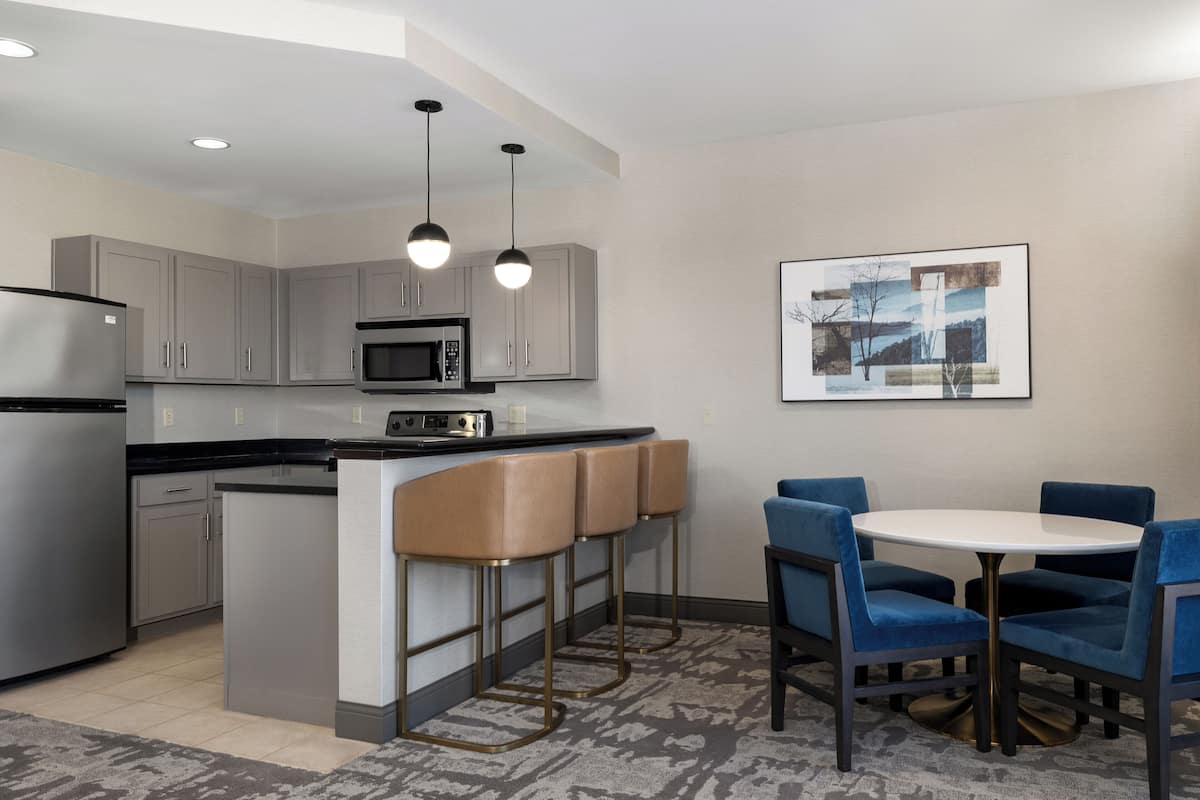 A modern kitchen with grey cabinets, a stainless steel refrigerator, a microwave, and a bar area with stools. There is a round table with blue chairs and a framed picture on the wall.