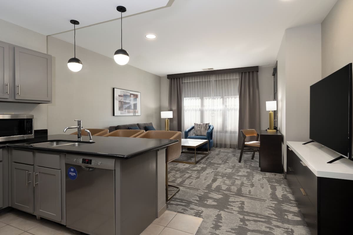 A modern hotel room with a kitchen area, a living area with a sofa and armchairs, a flat-screen TV, and a large window with curtains.