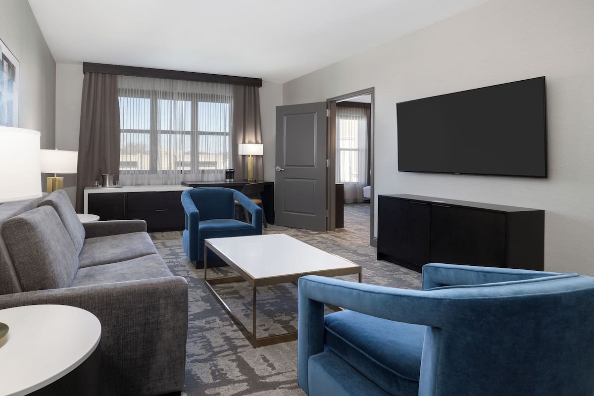 A modern hotel room with a grey sofa, blue armchairs, a small white table, a flat-screen TV mounted on the wall, and a large window with sheer curtains.