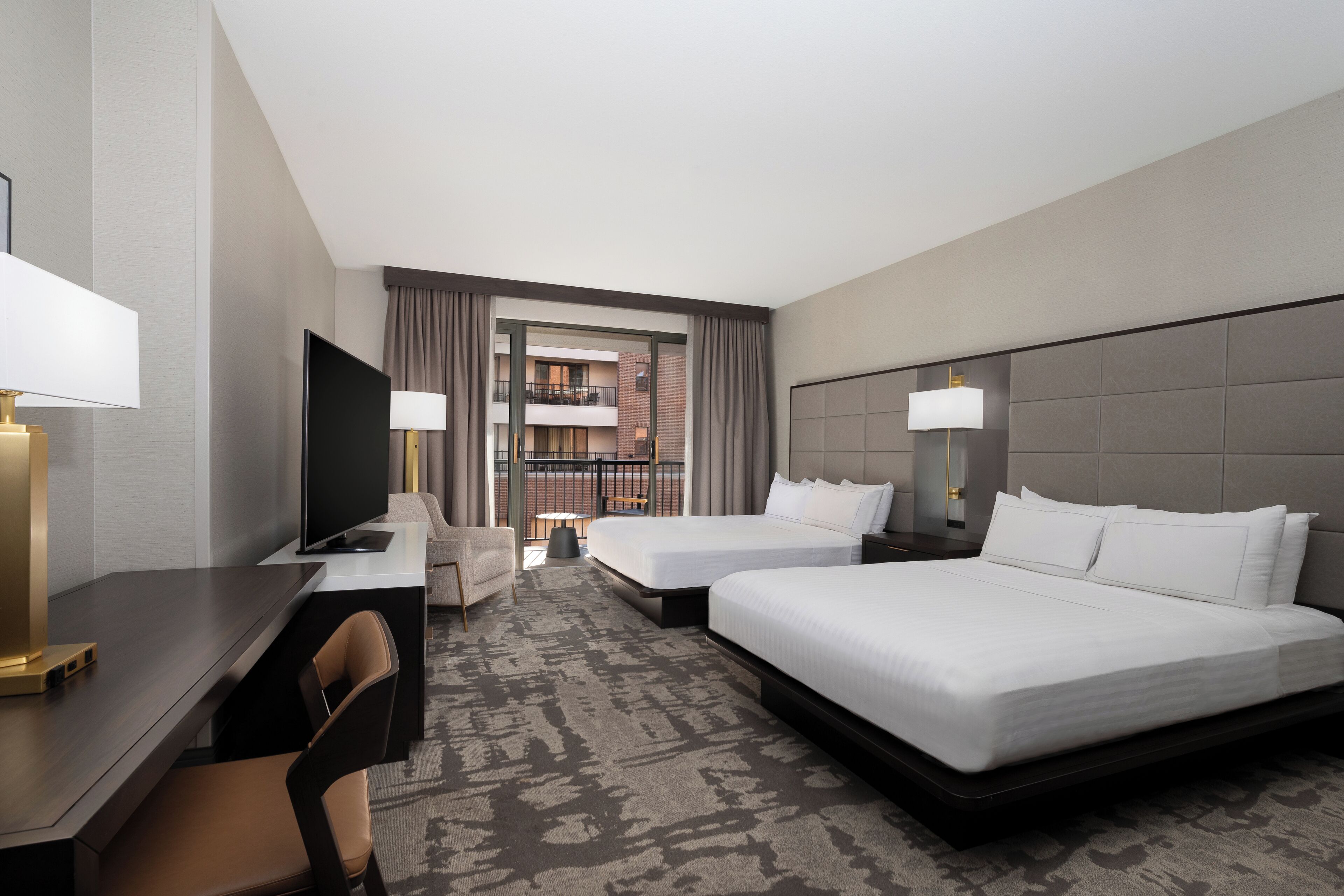 A hotel room with a desk, two beds, a TV, and a view of a building with balconies.