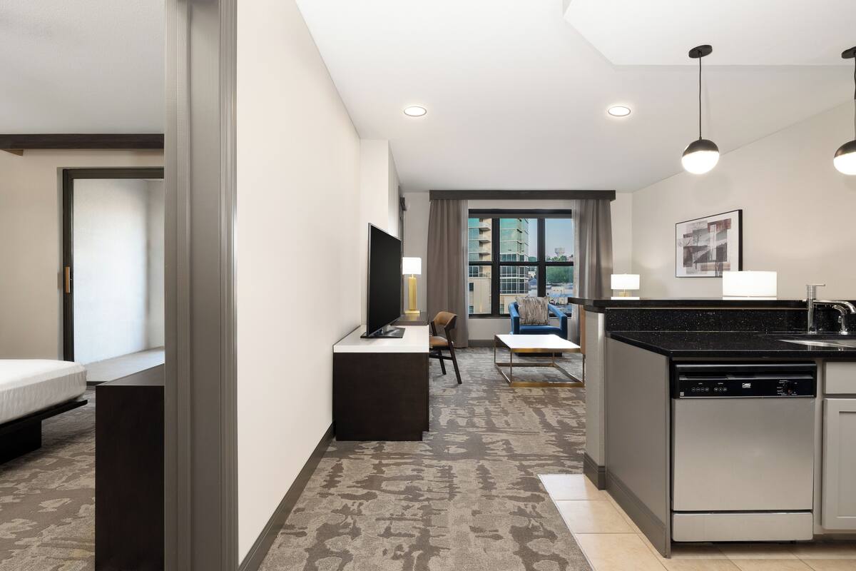 A modern hotel room with a kitchenette, a flat-screen TV, a sofa, and a dining area.