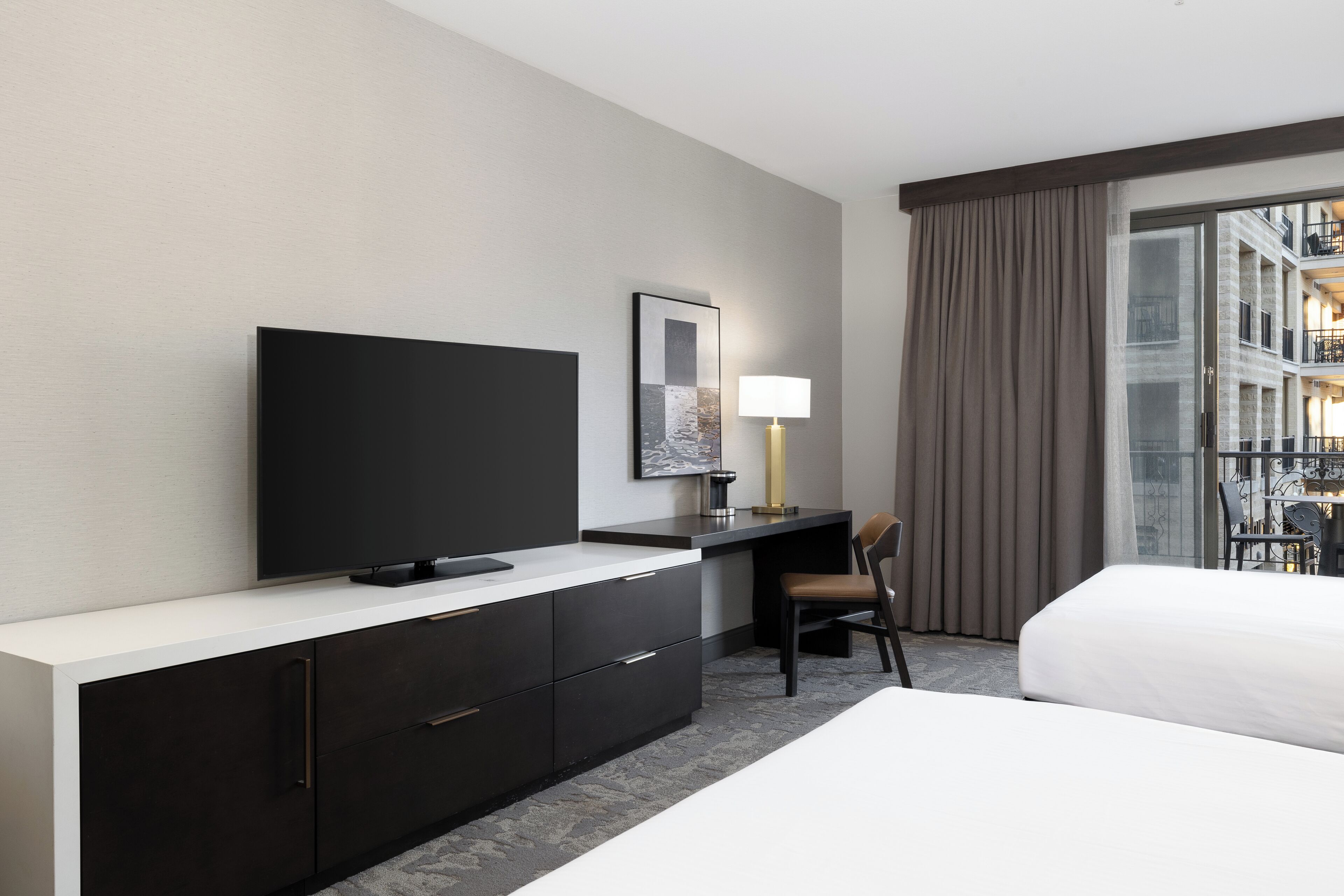 Una habitación de hotel con cama, escritorio, televisión, silla y ventana con cortinas.