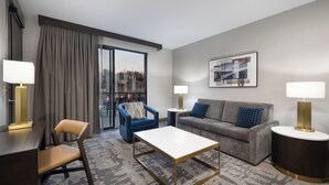 A modern hotel room with a grey sofa, a blue armchair, a wooden coffee table, and a large window with curtains.