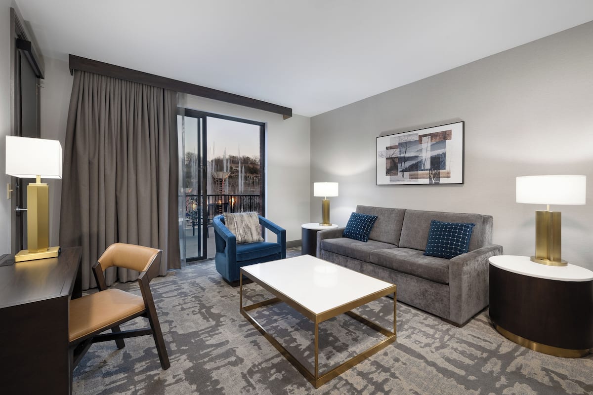 A modern hotel room with a grey sofa, a blue armchair, a wooden coffee table, and a large window with curtains.