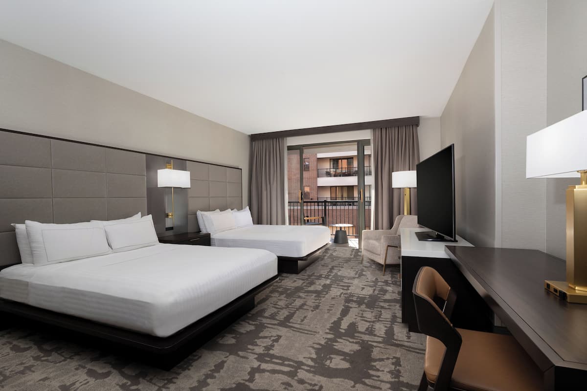 A hotel room with two beds, a desk, a chair, a TV, and a balcony with a view of a building.