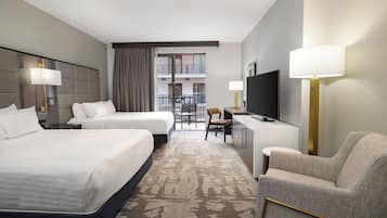 A hotel room with two beds, a desk, a chair, a TV, and a balcony with a view of buildings.