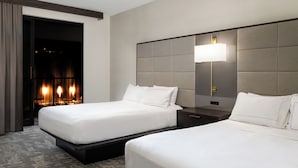 A hotel room with two beds, a nightstand, a lamp, and a view of a lit interior.