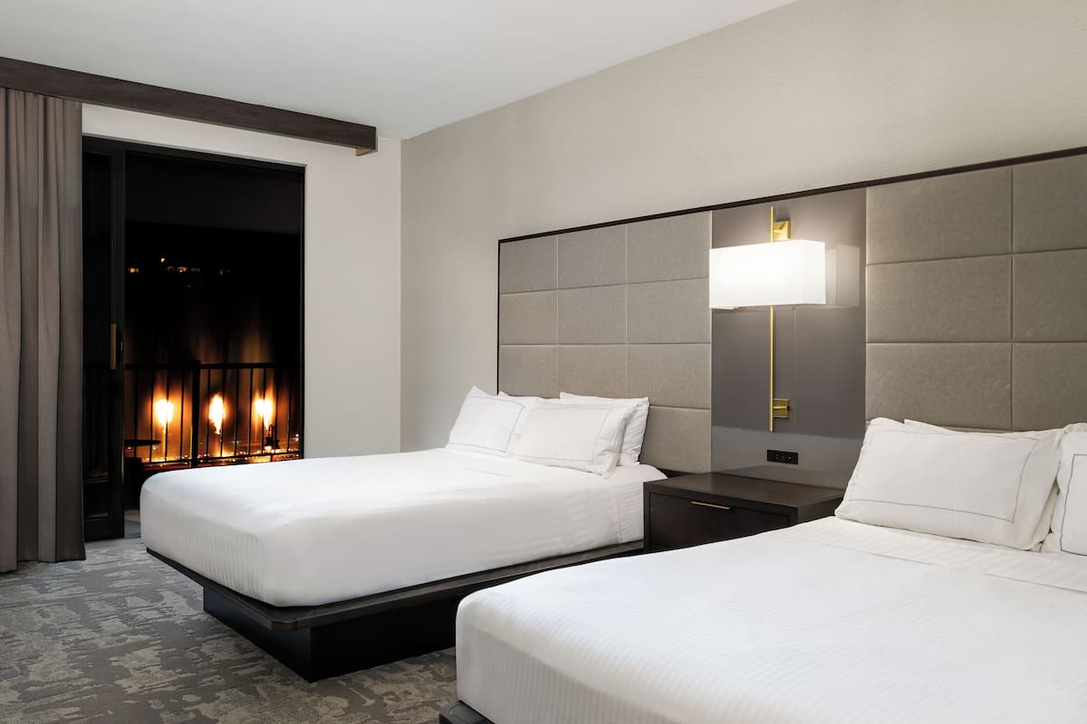 A hotel room with two beds, a nightstand, a lamp, and a view of a lit interior.