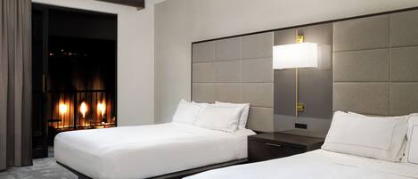 A hotel room with two beds, a nightstand, a lamp, and a view of a lit interior.