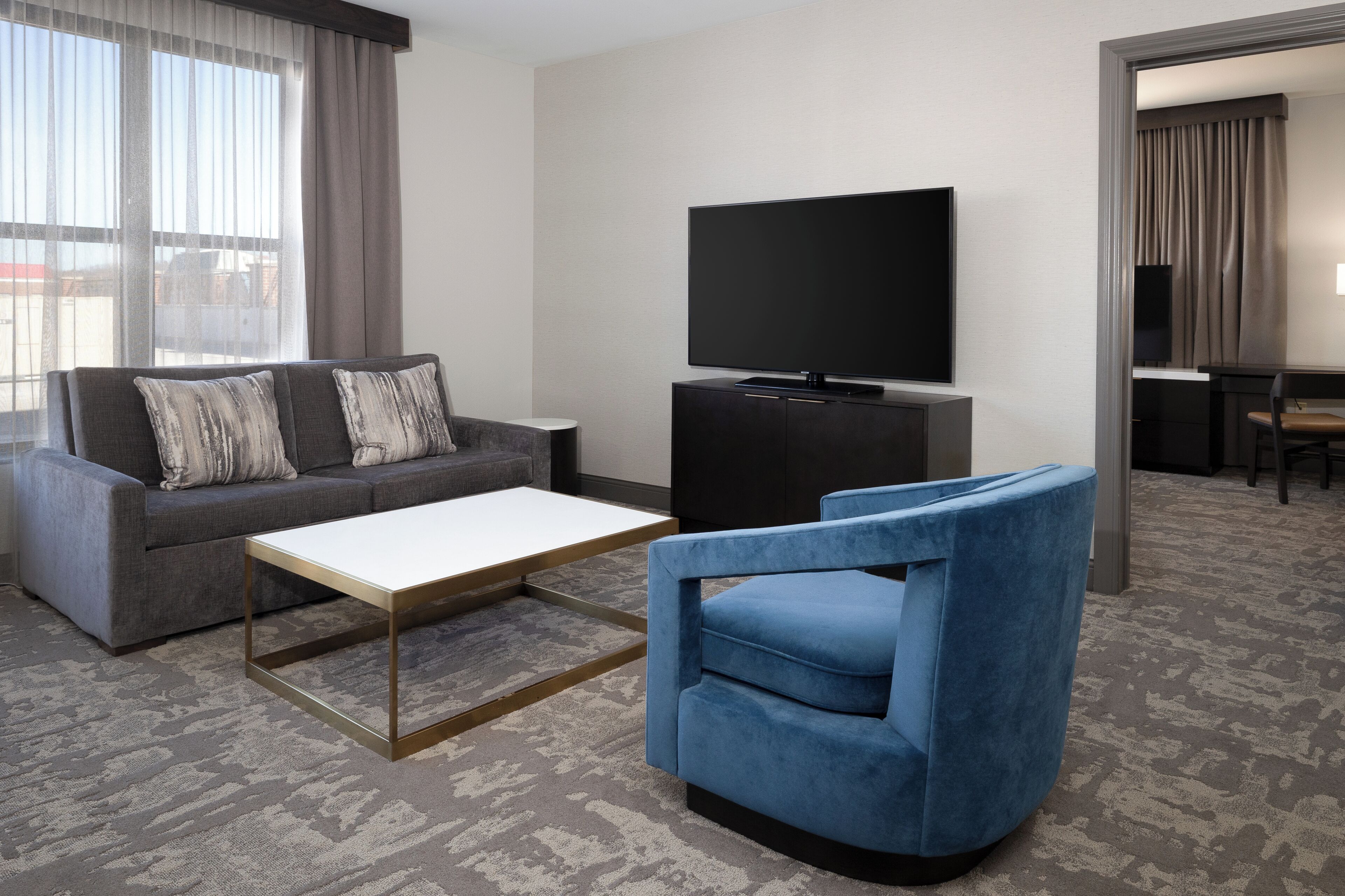 A modern hotel room with a grey sofa, a blue armchair, a flat-screen TV on a black stand, and a small table.