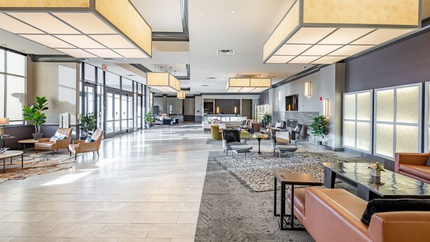 A spacious lobby with a modern design, featuring a central seating area with sofas and armchairs, a coffee table, and a fireplace. Large windows provide ample natural light, and the space is decorated with potted plants and a patterned rug.